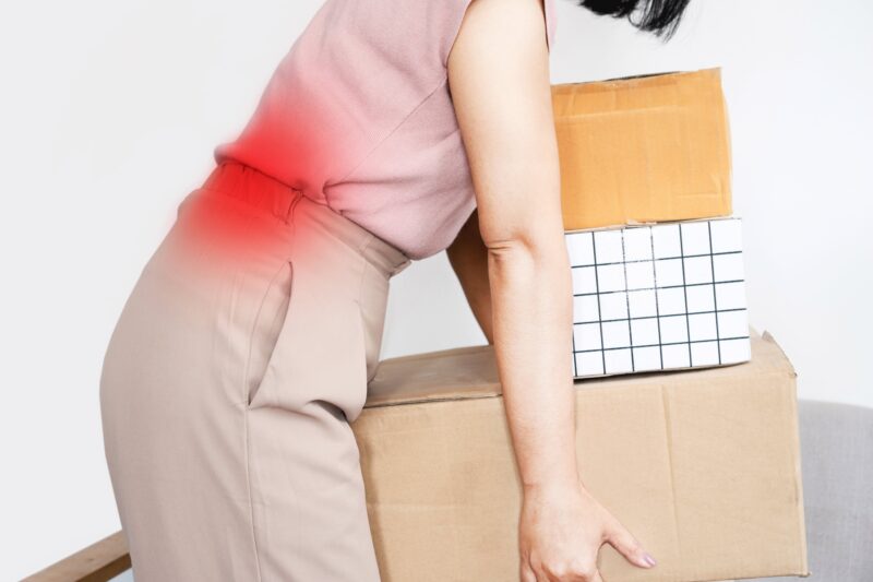 woman with a backache while lifting boxes, back pain caused by wrong movement or bad posture