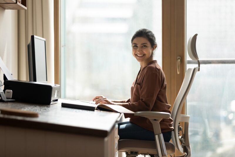 Portrait of happy biracial business woman freelancer sit by computer at comfy workplace at corporate workspace or at home
