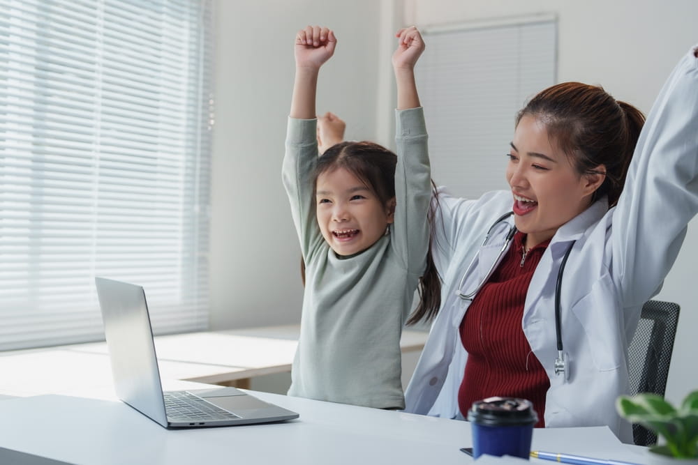 Asian female pediatrician and her young patient are raising arms in excitement while looking at laptop screen, celebrating successful treatment