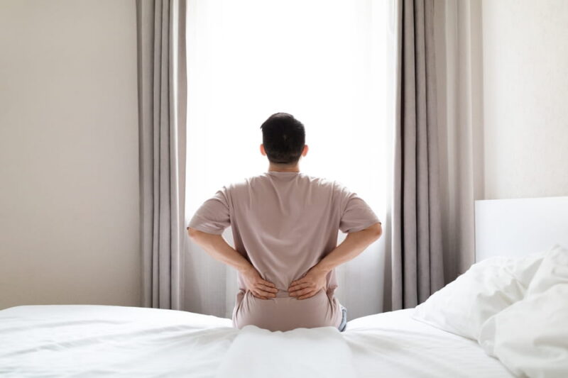 Tired man suffering from back pain after sleeping in bedroom, waking up in morning, sitting on bed and touching his lower back