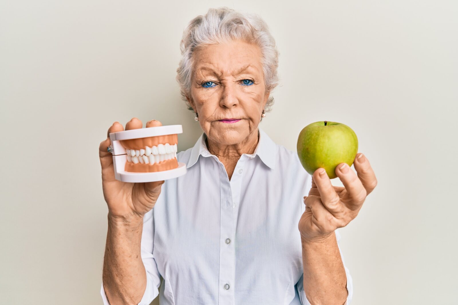 older lady holding dentures in one hand and an apple in the other hand with an unhappy expression