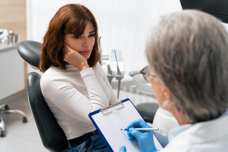 Young white brunette woman sits in the dentist's chair, holding onto her tooth in pain.