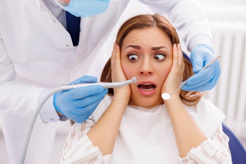 Woman afraid while sitting at dental chair at dentist office while doctor is holding dental drill and angled mirror, fixing patient's tooth