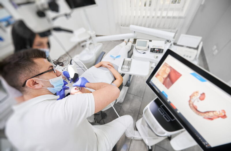 Dentist and assistant in medical masks using dental intraoral scanner while examining patient teeth in dental office.