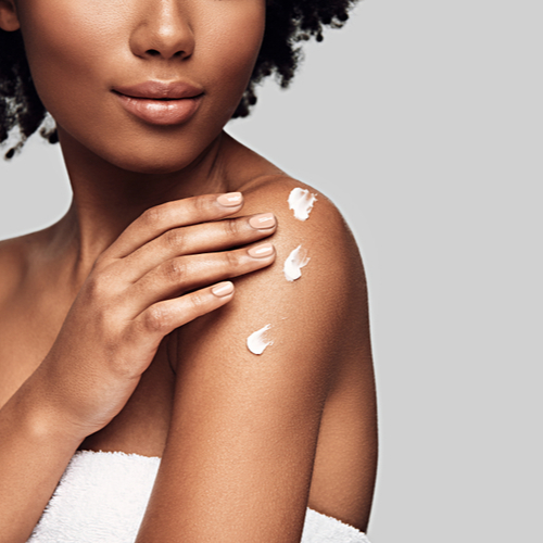 Beauty routine. Close up of young African woman applying body cream and smiling while standing against grey background