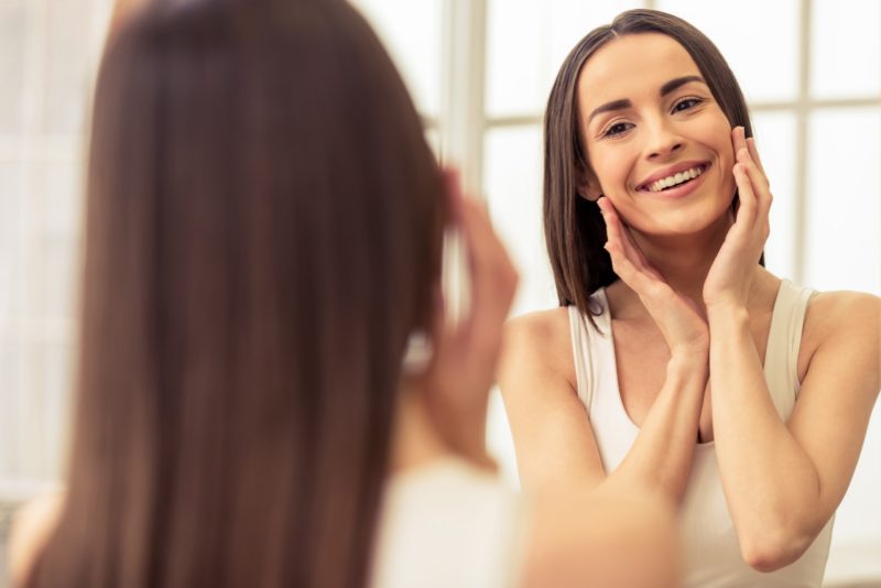 Young woman is touching her face and smiling while looking at the mirror
