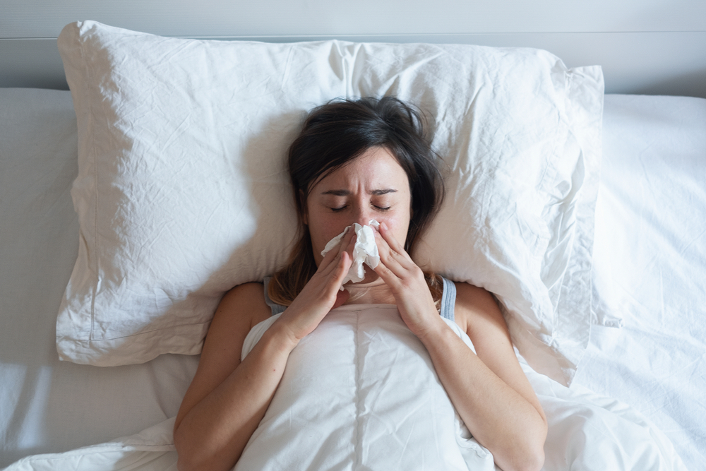 Young woman sneezing nose covered by warm duvet
