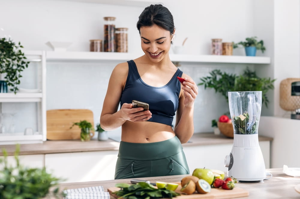 Shot of athletic woman using mobile phone while preparing smoothie in the kitchen at home.