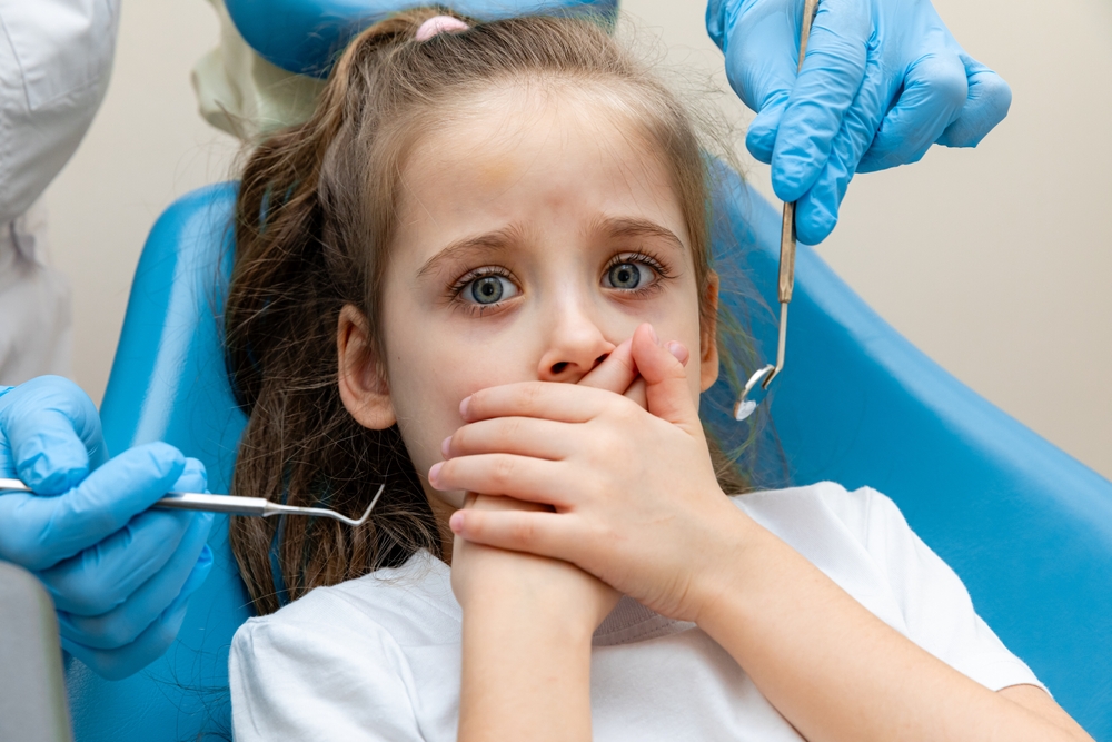 Cute little girl sitting in the dentist chair and covering her mouth in fear,