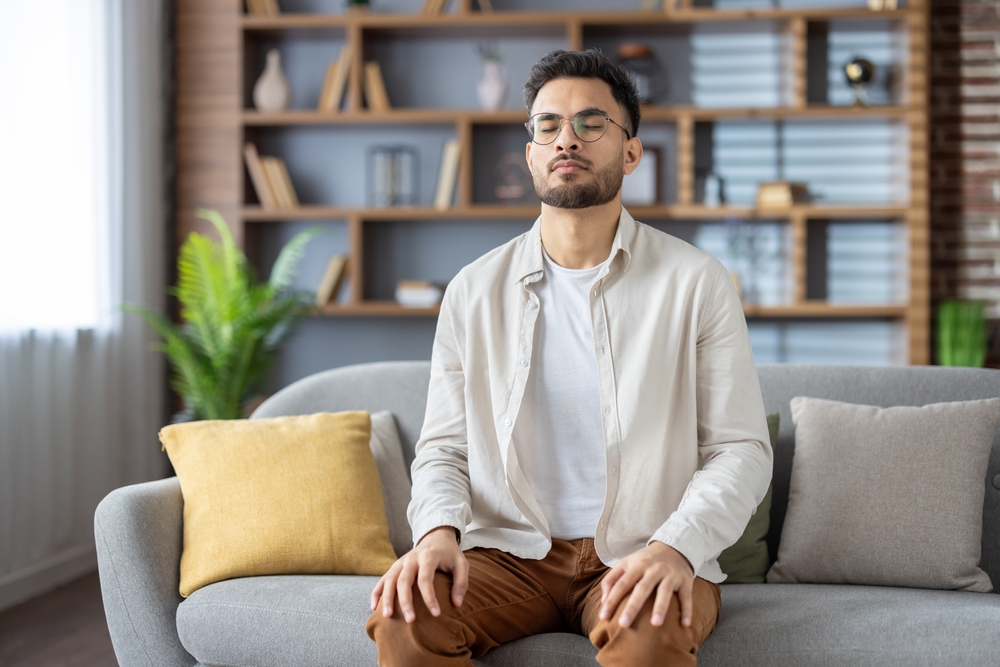 Young man sitting on sofa with eyes closed, practicing meditation and mindfulness