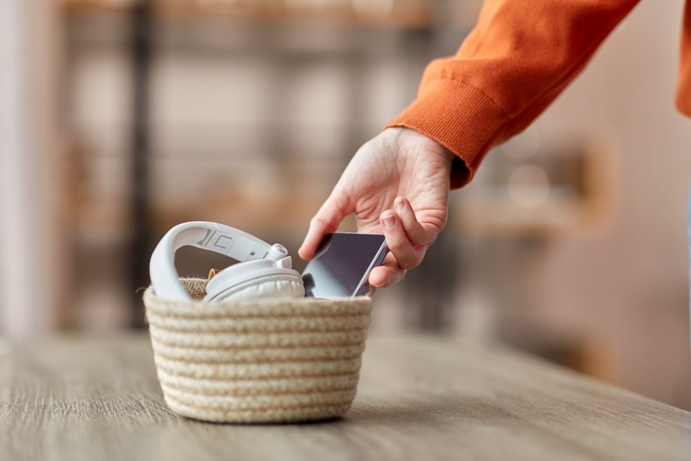 digital detox and technology concept - close up of hand holding smartphone and different gadgets in wicker basket on table at home