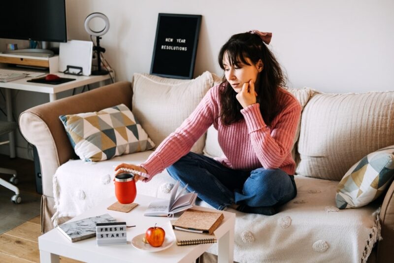Young adult woman sits cross-legged on sofa holding red coffee mug contemplating personal goals