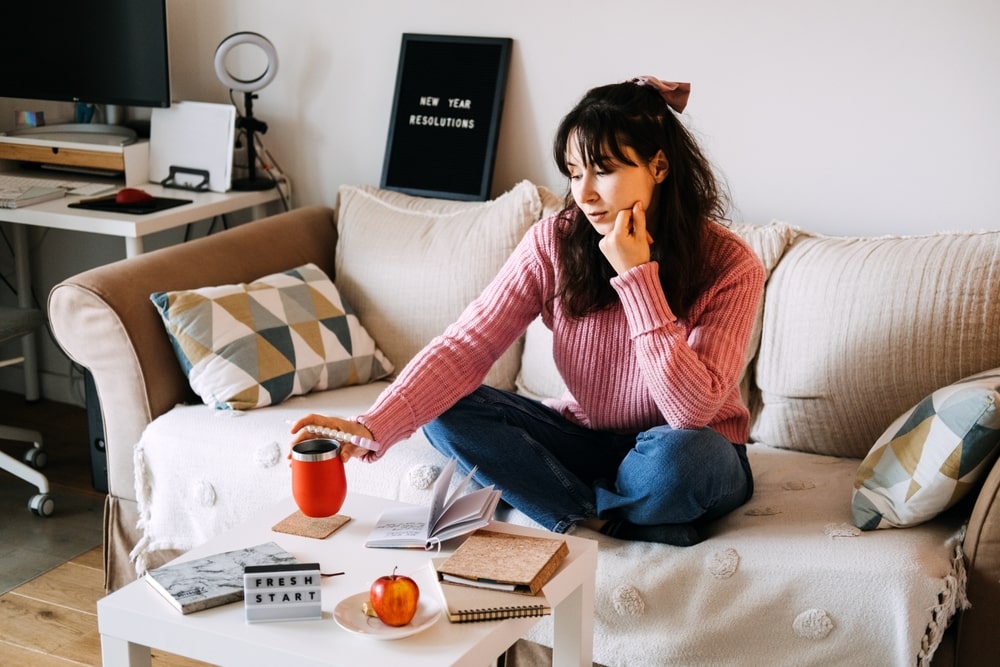 Young adult woman sits cross-legged on sofa holding red coffee mug contemplating personal goals