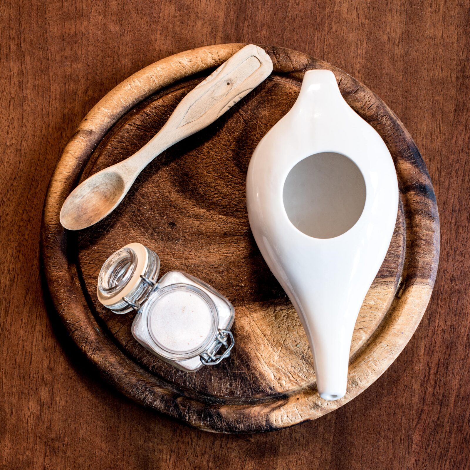 neti pot on a wood tray beside wooden spoon and jar of salt
