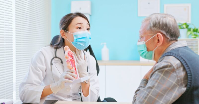 asian female doctor wearing face mask is showing ENT model and explaining to elder senior man patient in hospital