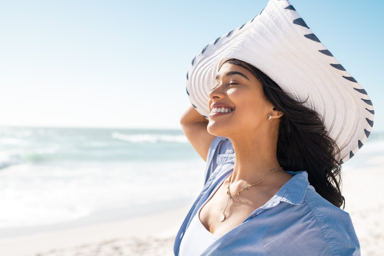 Beautiful latin hispanic woman wearing white straw hat at seashore with copy space. Young smiling woman on vacation enjoy sea breeze. Close up face of stylish woman wearing fashion straw hat at beach