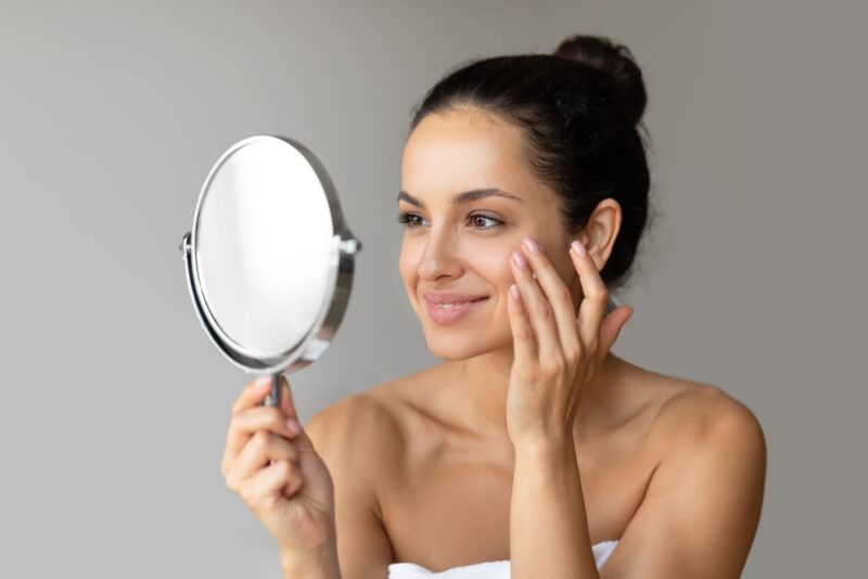 Attractive young woman wrapped in towel applying face cream, looking at round mirror and smiling
