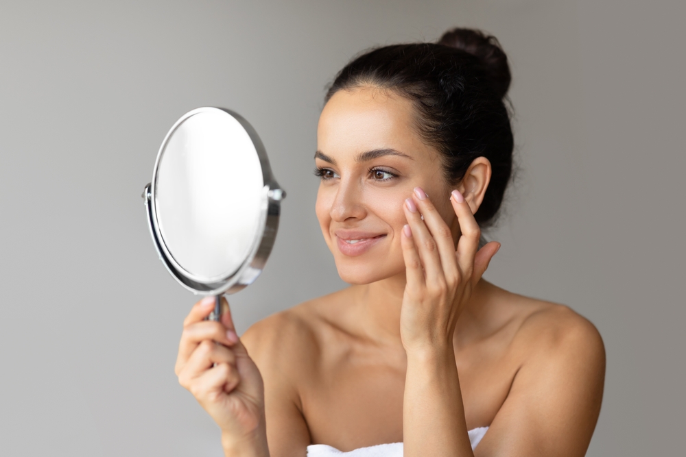 Attractive young woman wrapped in towel applying face cream, looking at round mirror and smiling
