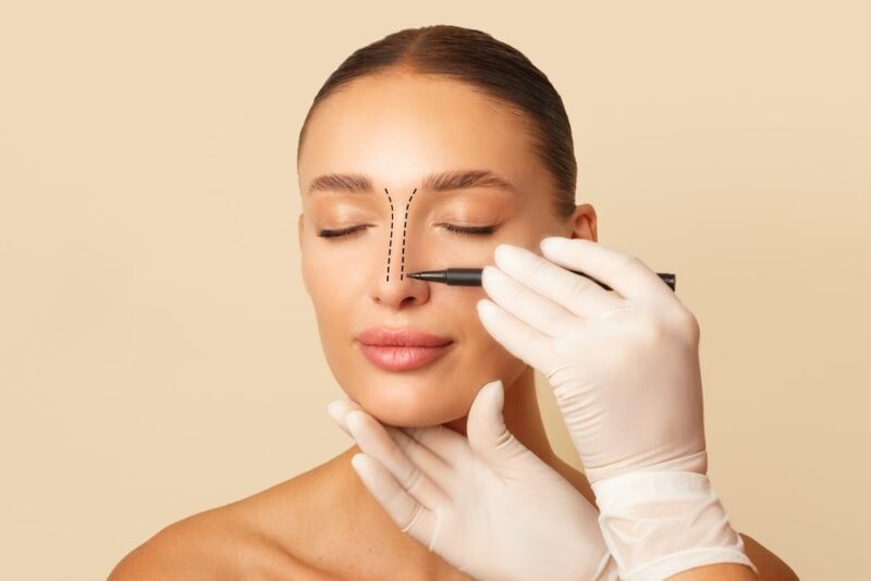 Doctor making marks on female nose, young woman posing on beige studio background with closed eyes during consultatio