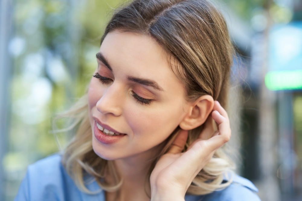 Close up portrait of young woman, tuck hair behind ear, looking flirty and smiling, sitting in blue shirt outdoors on street.
