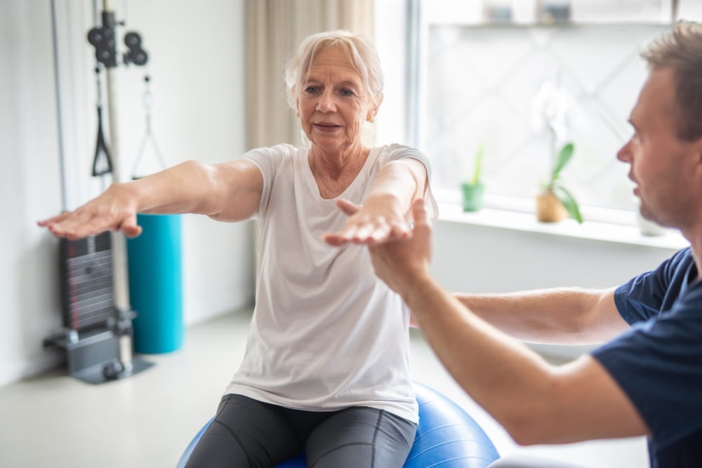 Senior woman doing physical therapy exercises with physiotherapist on swiss ball.