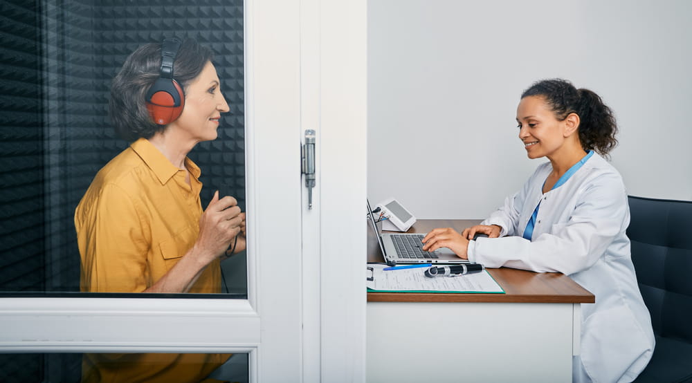 Audiological exam. Audiologist doing hearing check-up to a senior citizen woman in soundproof audiometric booth. Hearing loss treatment, side view