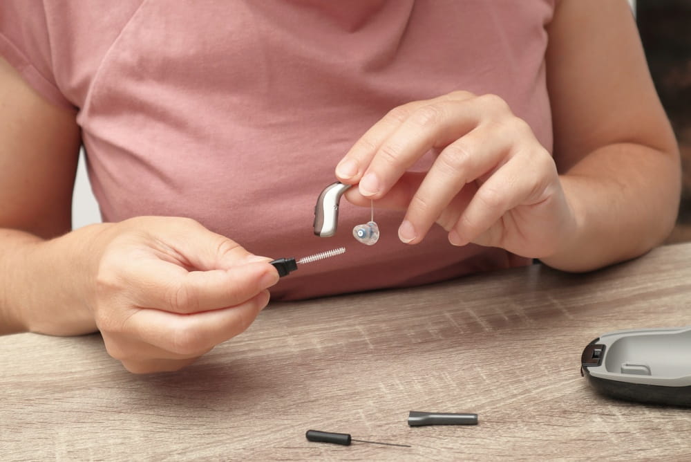 A close-up of a person's hands using a specialized cleaning brush to maintain a behind-the-ear hearing aid device for better performance and hygiene.