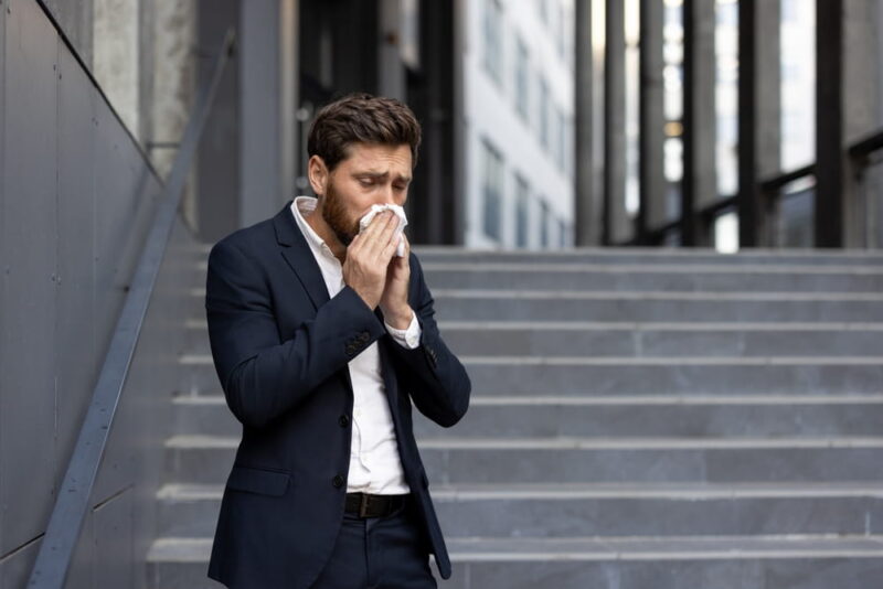 Caucasian manager sneezing and blowing his nose with a tissue while standing on office stairs, experiencing the discomfort of seasonal influenza or a cold in a corporate environment