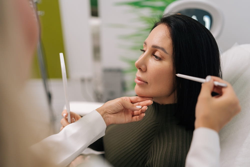 Side view of woman looking in mirror while cosmetologist marking face for aesthetic procedure, preparing for beauty treatment in cosmetology clinic.