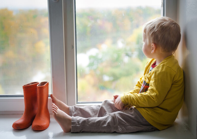 Adorable little blond kid boy sitting on the windowsill with rain shoes and looking on autumn raindrops