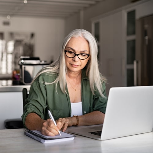 Senior stylish woman taking notes in notebook while using laptop