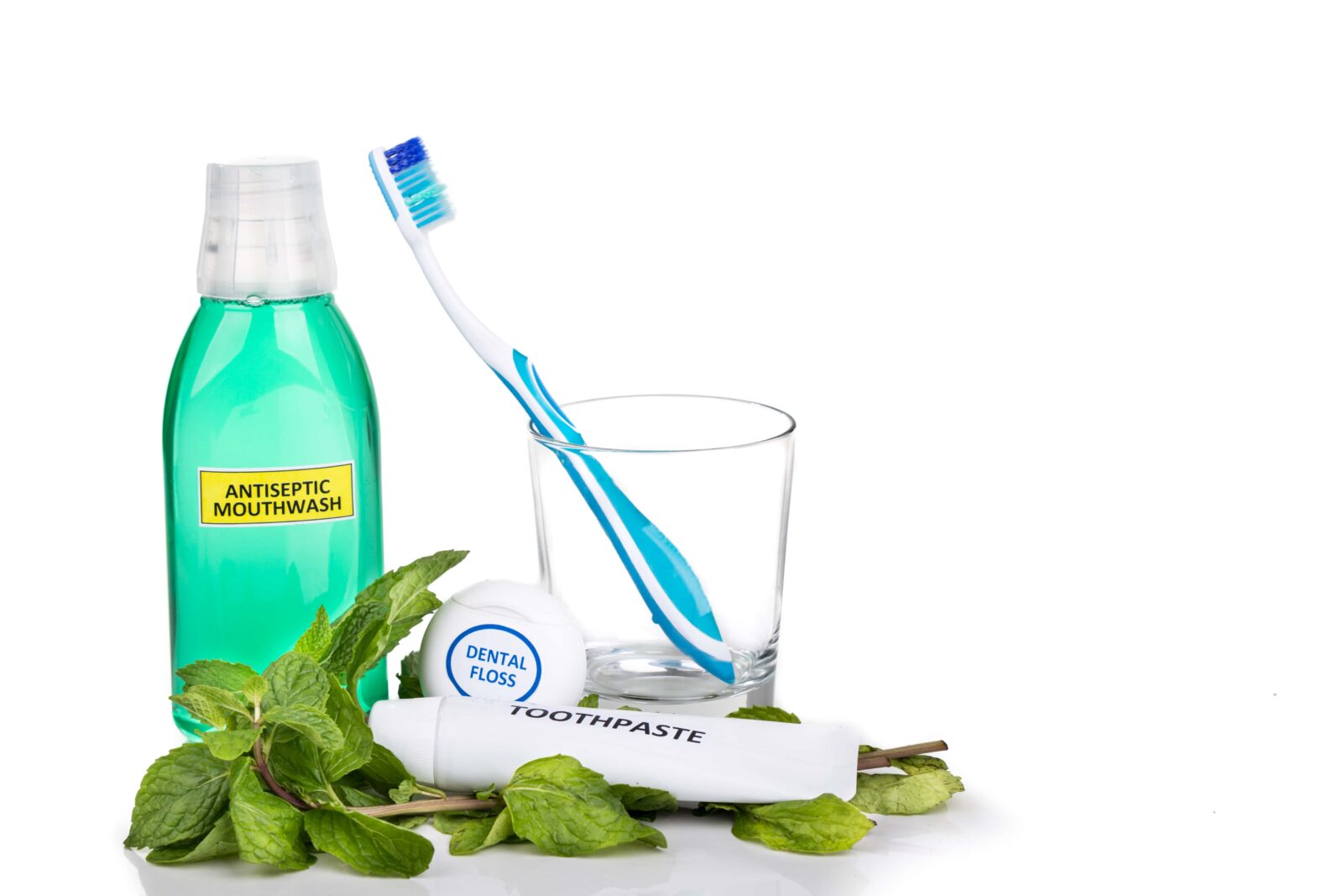 assorted dental care tools: mouthwash, toothbrush in a cup, dental floss, and toothpaste all surrounded by mint leaves on a white background