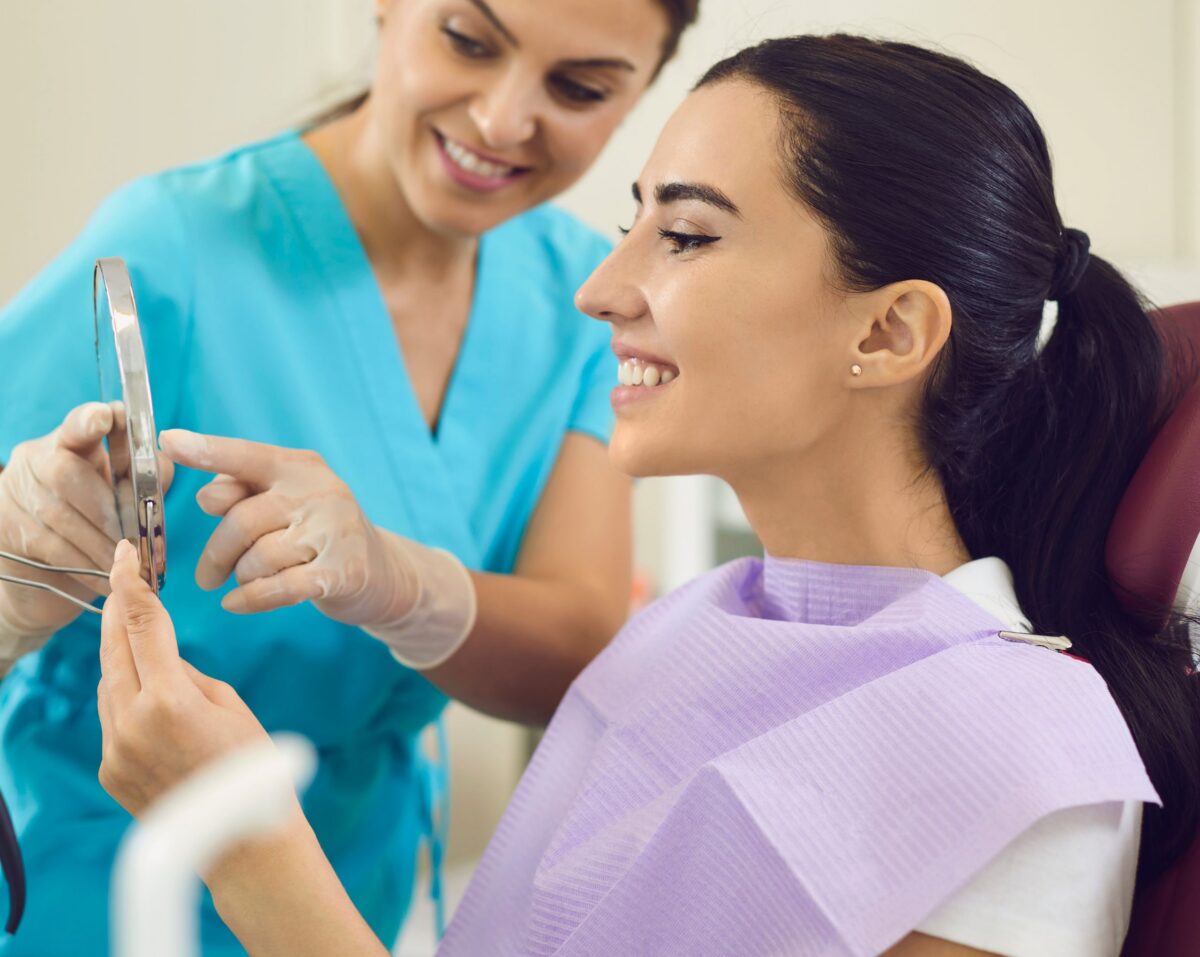 Young, female dental patient admiring smile in a mirror as dental assistant looks on