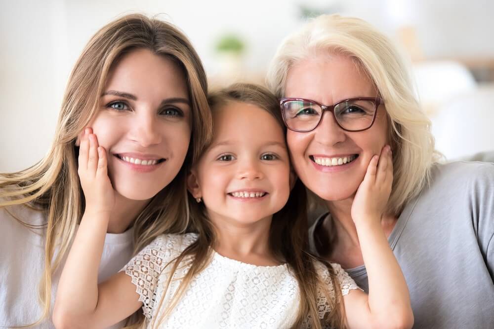 A young girl, her mother, and her grandmother smiling together with whitened teeth.
