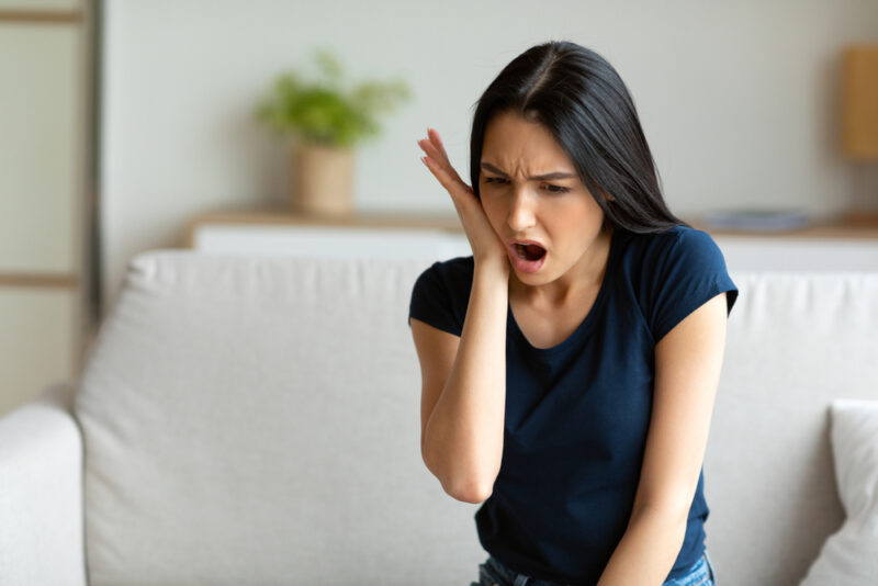 Toothache. Girl Suffering From Tooth Pain Touching Cheek Sitting On Couch At Home. Dental Problem Concept.