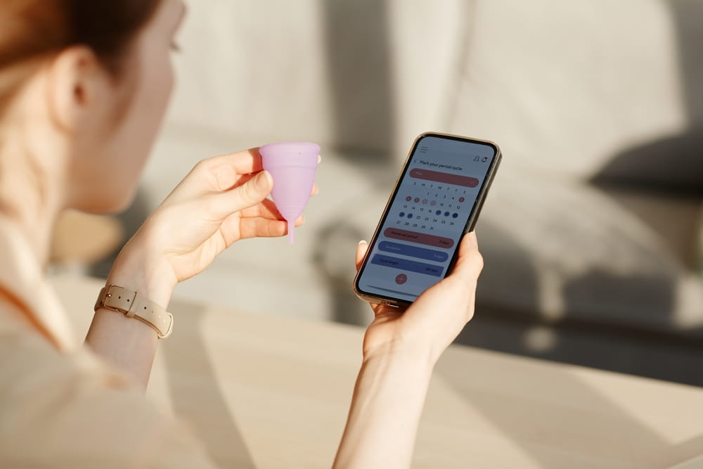 Close up of young woman calculating menstrual cycle using mobile app and holding reusable cup