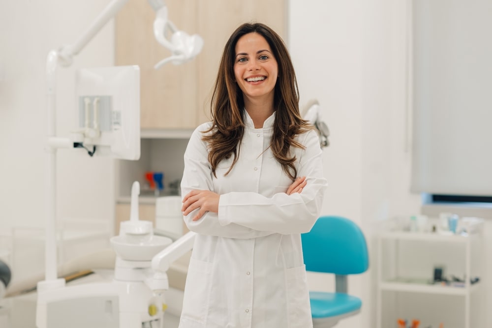 Confident female dentist smiling with crossed arms, standing in her modern dental clinic