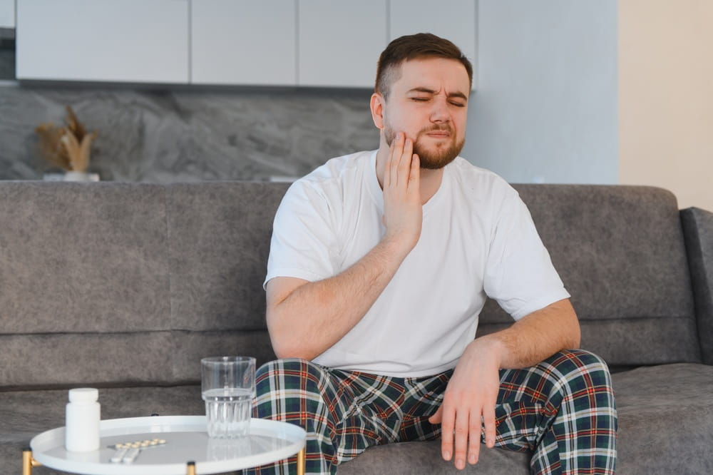 Young man grimacing on couch holding cheek in pain, needing medication for severe toothache and oral inflammation at home