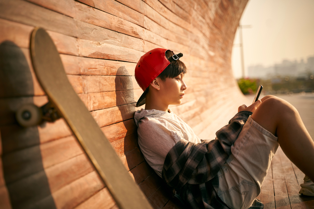teenage asian skateboarder boy looking at cellphone while resting