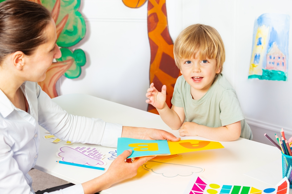 Early development lesson, woman shows and teaches letters to little blond boy looking at camera