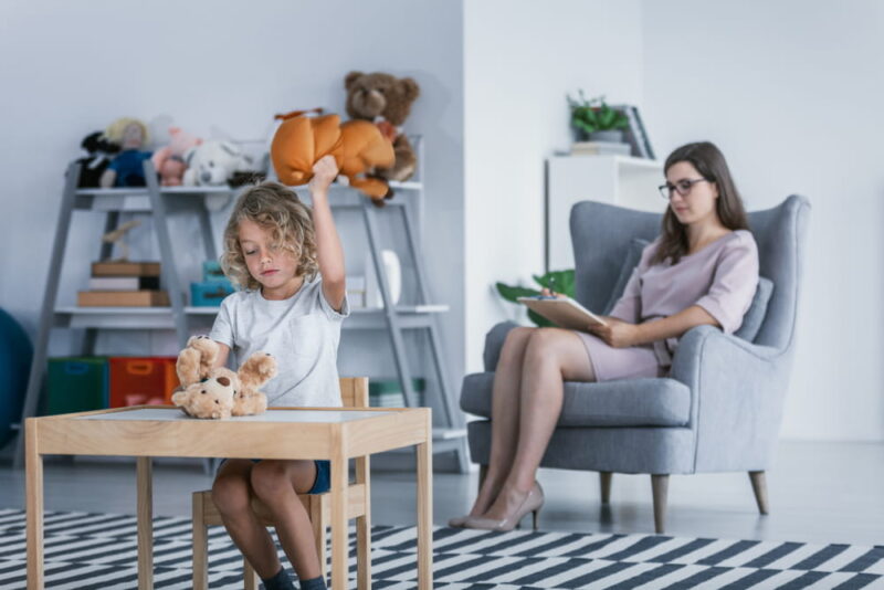 A child with behavioral problems sitting by a table and hitting a teddy bear during a therapeutic meeting with a therapist