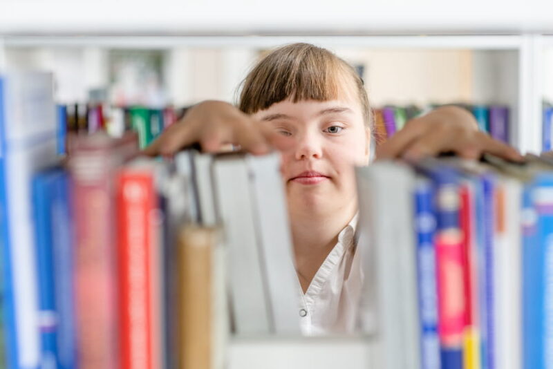 Girl with syndrome down chooses a book on a shelf in the library. Education for disabled children concept