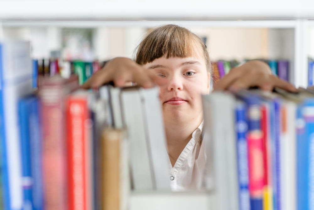 Girl with syndrome down chooses a book on a shelf in the library. Education for disabled children concept