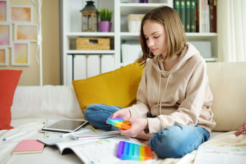 Teenage girl playing with rainbow pop-it fidget toy while studying at home