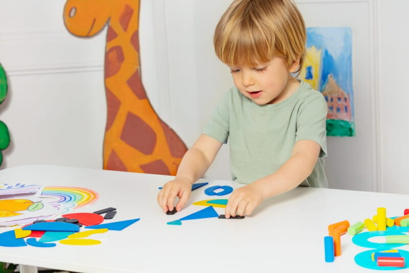 Young little blond boy form shapes standing by the table in the kindergarten playing developmental games