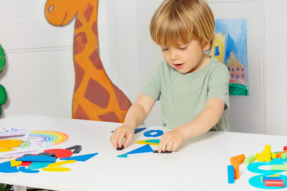 Young little blond boy form shapes standing by the table in the kindergarten playing developmental games