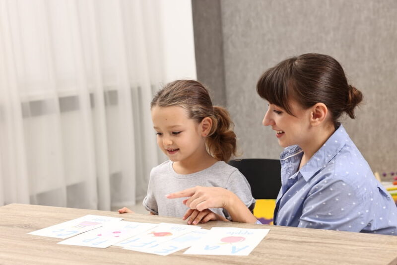 Smiling speech therapist working with little girl at table in autism treatment center. Space for text