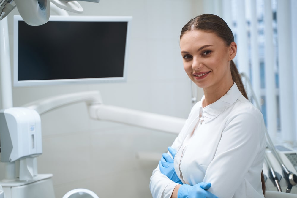 Smiling woman in white uniform and sterile gloves is standing in modern dental clinic near digital screen