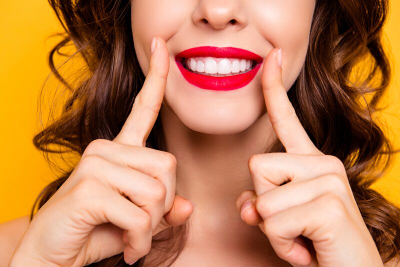 Close up cropped half face portrait of cheerful toothy woman with white teeth making surgery examination measuring standard size of smile with forefingers. Correction operation contouring concept