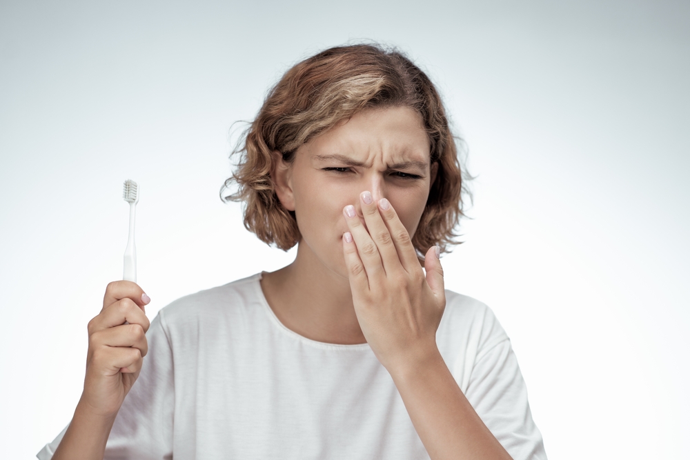 A woman in a white t-shirt covers her mouth and holds a toothbrush, showing signs of discomfort.